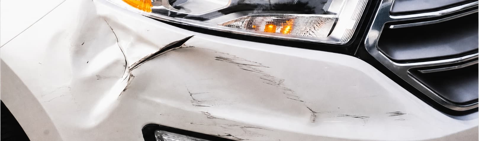 Close-up of a white SUV's front fender, crumpled near the headlight with scrape marks across the bumper